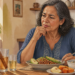 Mujer adulta sentada a la mesa frente a un plato con legumbres y aguacate, junto a una infusión con ramas de canela, un pequeño vaso de bebida clara, avena con frutos rojos, fruta y un teléfono móvil.