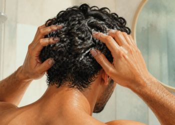 Hombre en la ducha masajea el cuero cabelludo con champú mientras se lava el pelo, con espuma visible y un espejo al fondo.