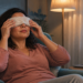 Mujer sentada en un sillón coloca una compresa de tela sobre los ojos en una sala iluminada por una lámpara, con un portátil y un cuenco sobre una mesa auxiliar.