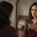 Mujer adulta observa un cepillo para el cabello frente al espejo, con expresión de preocupación, en un dormitorio iluminado.