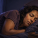 Mujer dormida de lado en una cama durante la noche, con una férula dental transparente en un estuche abierto sobre la mesilla junto a una lámpara encendida.