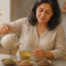 Mujer adulta vierte agua caliente de una tetera en una taza de vidrio con semillas, junto a tres pequeños platos con distintas semillas sobre la encimera de una cocina.