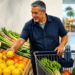 Hombre adulto selecciona un limón junto a un carrito con verduras, sandía y una botella reutilizable en la sección de frutas y hortalizas de un mercado.