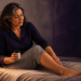 Mujer adulta sentada en la cama por la noche sostiene un frasco de suplemento mientras se toca una pierna, junto a un vaso de agua sobre la mesita.
