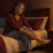 Mujer adulta sentada en la cama por la noche se sujeta la pierna y el pie, con una lámpara encendida y dos vasos sobre la mesita de noche.