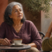 Dos mujeres mayores conversan sentadas frente a frente en una mesa al aire libre, una de ellas con una taza delante y expresión atenta durante la conversación.