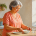 Mujer mayor en una cocina colocando un cuenco con un alimento blanco sobre la mesa, junto a un plato y un vaso de agua.