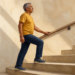 Hombre mayor subiendo escaleras con una mano apoyada en la barandilla, vestido con camiseta amarilla, pantalón azul y zapatillas deportivas.