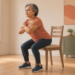 Mujer mayor realiza una sentadilla asistida frente a una silla en una habitación luminosa, como ejercicio en casa para fortalecer caderas y glúteos después de los 60.