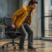 Hombre de pie junto a una silla de oficina, incorporándose con gesto de molestia en las piernas en un espacio de trabajo con escritorio, portátil y ventanales.