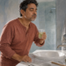 Hombre de mediana edad haciendo un enjuague bucal frente al espejo del baño mientras sostiene un vaso con líquido claro, junto a un pequeño recipiente y una botella.