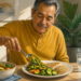 Hombre mayor sentado a la mesa mientras sirve verduras en su plato con una cuchara, acompañado de pescado, un cereal y un vaso de agua en un comedor luminoso.