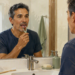 Hombre adulto frente al espejo del baño limpiándose la lengua con un raspador lingual, junto a un vaso de agua, un cepillo de dientes, jabón y dispensadores sobre el lavabo.