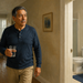 Hombre adulto de pie en un pasillo de su casa sosteniendo un vaso de agua, junto a la entrada de un baño visible al fondo.