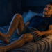 Hombre recostado en la cama por la noche mueve las piernas con gesto de incomodidad, junto a una lámpara encendida en la mesita de noche.
