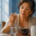 Mujer de mediana edad observa una pastilla blanca que sostiene con la mano mientras está sentada ante un vaso de agua, una taza de café y un frasco blanco.