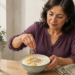 Mujer preparando un cuenco con una base cremosa, rodajas de plátano, avena y semillas, acompañado por un vaso de agua en una cocina luminosa.