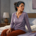 Mujer sentada en la cama al despertar, girando el cuello con gesto de dolor en un dormitorio luminoso, en una escena relacionada con el dolor de cuello matutino.