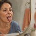 Mujer adulta frente al espejo del baño sacando la lengua para observarla, con un lavabo, una toalla y pequeños elementos decorativos visibles alrededor.