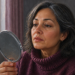 Mujer adulta observando sus labios en un espejo de mano, con sequedad visible alrededor de la boca, dentro de una habitación iluminada por una ventana.