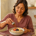 Mujer adulta en la cocina espolvoreando un ingrediente sobre un cuenco de avena con fruta, junto a un frasco de copos de avena y una manzana.