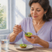 Mujer comiendo medio kiwi verde con una cuchara en una cocina luminosa, con otro medio kiwi sobre un plato y un vaso de agua en la mesa.