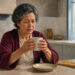 Mujer mayor sentada a la mesa de la cocina sostiene una taza caliente con ambas manos y observa sus dedos con gesto de incomodidad durante la mañana.