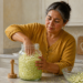 Mujer preparando col rallada en un frasco de vidrio para fermentarla, con un cuenco de col y un utensilio de madera sobre la encimera de una cocina.