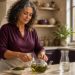Mujer madura prepara una infusión en una tetera de vidrio sobre la encimera de una cocina, junto a hierbas secas, una jarra de agua y una lata de bebida.