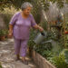 Mujer mayor regando con una regadera metálica un huerto doméstico con plantas y tomates, mientras camina por un jardín exterior.