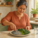 Mujer de mediana edad prepara un plato con pescado pequeño y verduras verdes en una cocina luminosa, junto a un vaso de agua sobre la encimera.
