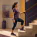 Mujer de mediana edad sube un escalón apoyándose en la barandilla, con ropa deportiva y zapatillas, dentro de una vivienda luminosa.