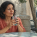 Mujer adulta sosteniendo un vaso de agua con ambas manos mientras está sentada en una terraza exterior, con pequeños frascos sobre una mesa al fondo.