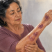 Mujer mayor observa varios moretones de color morado en el antebrazo mientras sostiene el brazo con la otra mano, junto a una ventana iluminada.