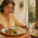 Mujer adulta sentada a la mesa comiendo un plato con pescado, verduras y arroz, con un vaso de agua delante y una fuente de fritos al fondo.