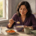 Mujer sentada a la mesa come una preparación de cereal cocido junto a un plato de verduras, una taza y un vaso de agua en una cocina luminosa.