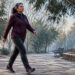 Mujer de mediana edad caminando a paso ligero por un sendero de parque arbolado, vestida con ropa deportiva y zapatillas.