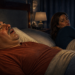 Hombre acostado boca arriba en la cama con la boca abierta mientras ronca, junto a una mujer despierta a su lado que lo mira con molestia en un dormitorio nocturno.