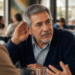 Hombre mayor en una cafetería inclinándose hacia dos personas y acercando una mano al oído para escuchar mejor durante una conversación, en una escena relacionada con la audición con la edad.