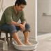Hombre sentado en un baño con los pies dentro de una palangana con agua, junto a una botella transparente, en una escena relacionada con un baño casero para el olor de pies.