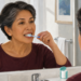 Mujer mayor cepillándose los dientes frente al espejo del baño, con un recipiente de hilo dental junto al lavabo durante su rutina de higiene bucal.