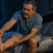 Hombre mayor sentado en la cama durante la noche, sujetándose la pantorrilla y el pie con gesto de dolor, junto a un vaso de agua sobre la mesita.