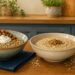 Dos cuencos de avena sobre una mesa de cocina, uno con fruta, yogur y frutos secos, y otro más simple junto a un paquete abierto con copos de avena derramados.