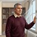 Hombre mayor abre una ventana en una habitación de casa mientras la cortina se mueve con el aire, junto a un radiador y una estantería al fondo.