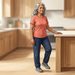 Mujer mayor de cabello gris realiza un ejercicio de equilibrio en la cocina, apoyando una mano sobre la encimera mientras mantiene una pierna ligeramente elevada.