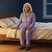 Mujer mayor sentada en el borde de la cama durante la noche, con pijama lila y un pequeño punto de luz encendido junto a la puerta del dormitorio.