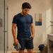 Hombre adulto de pie en un baño, vestido con camiseta y pantalón corto, sosteniendo una toalla junto a una ducha y un lavabo.