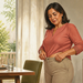 Mujer adulta de pie en el comedor ajustándose la cintura del pantalón después de comer, junto a una mesa con platos, un vaso de agua y un ambiente doméstico luminoso.