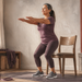 Mujer adulta haciendo una sentadilla frente a una silla en casa, con ropa deportiva, mancuernas y una esterilla de ejercicio alrededor.