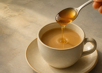 Cuchara con miel vertiéndose sobre una taza de bebida caliente de color claro colocada sobre un plato.